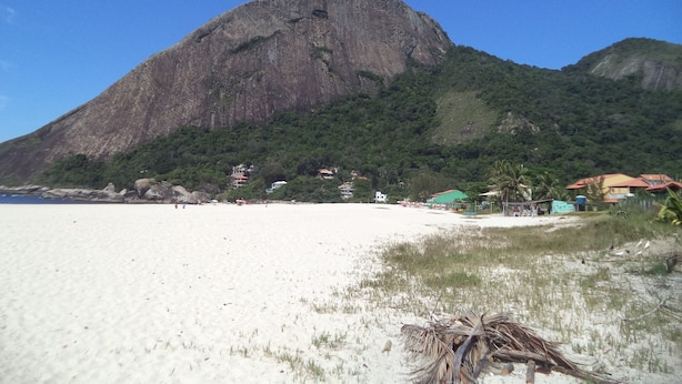 Ein Strand mit einem großen Berg im Hintergrund, einem sandigen Ufer und einigen Gebäuden nahe am Wasser.