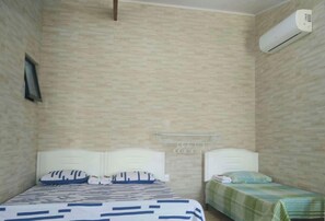 Une chambre avec deux lits, l’un recouvert d’une couvre-lit à rayures et l’autre d’une couverture à carreaux verts. On y trouve un climatiseur fixé au mur et une petite étagère avec des objets suspendus.