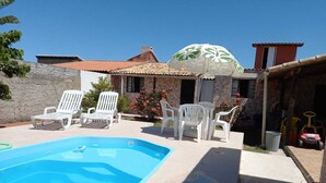 Ein Swimmingpool mit weißen Liegestühlen, einer überdachten Terrasse und einem Haus mit einem Ziegeldach.