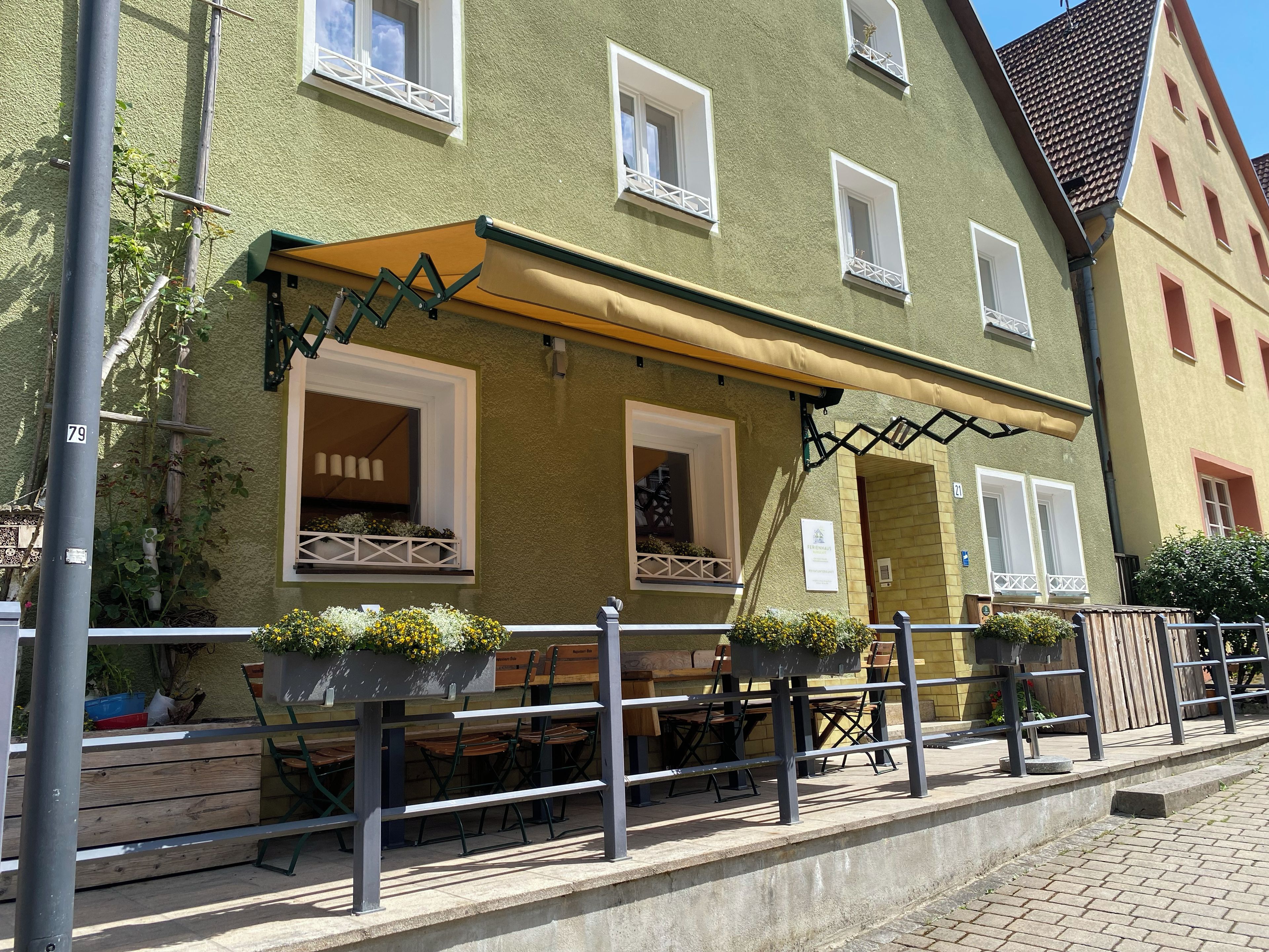 A building with a green facade, multiple windows, outdoor seating with tables and chairs, and a yellow awning.