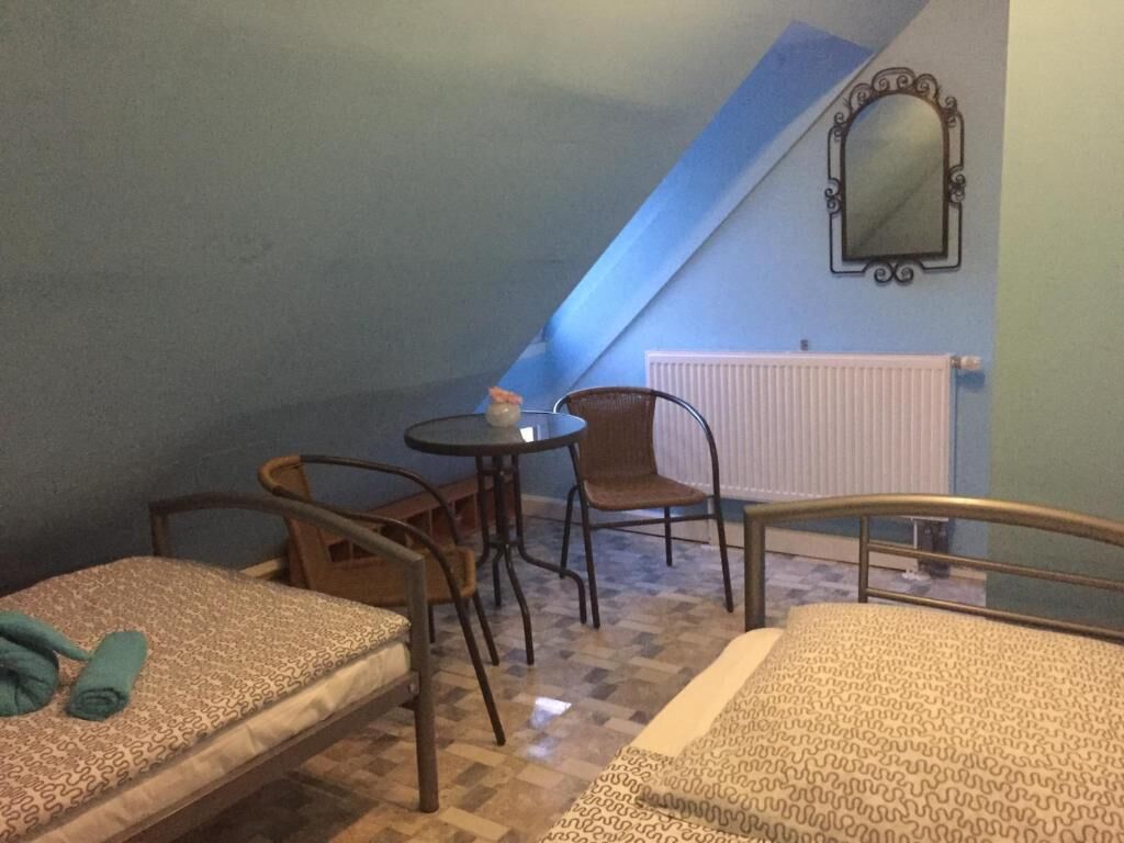 A small, cozy room with two beds, a table, and a chair. There is a window with a view of the sky and a radiator on the wall.