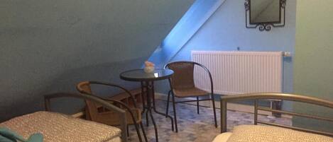 A small, cozy room with two beds, a table, and a chair. There is a window with a view of the sky and a radiator on the wall.