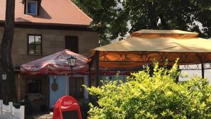 Un bâtiment avec une boîte aux lettres rouge au logo Coca-Cola, des tables et des chaises à l’extérieur, protégées par des parasols, et un grand buisson au premier plan.