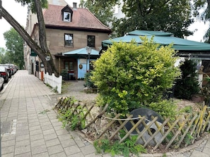 Un edificio con techo de tejas, acera pavimentada y un pequeño jardín con cerca de madera y arbusto.