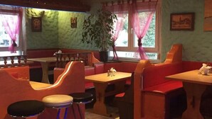 Un restaurant avec des banquettes et des tabourets hauts, une grande plante en pot et une fenêtre avec des rideaux.