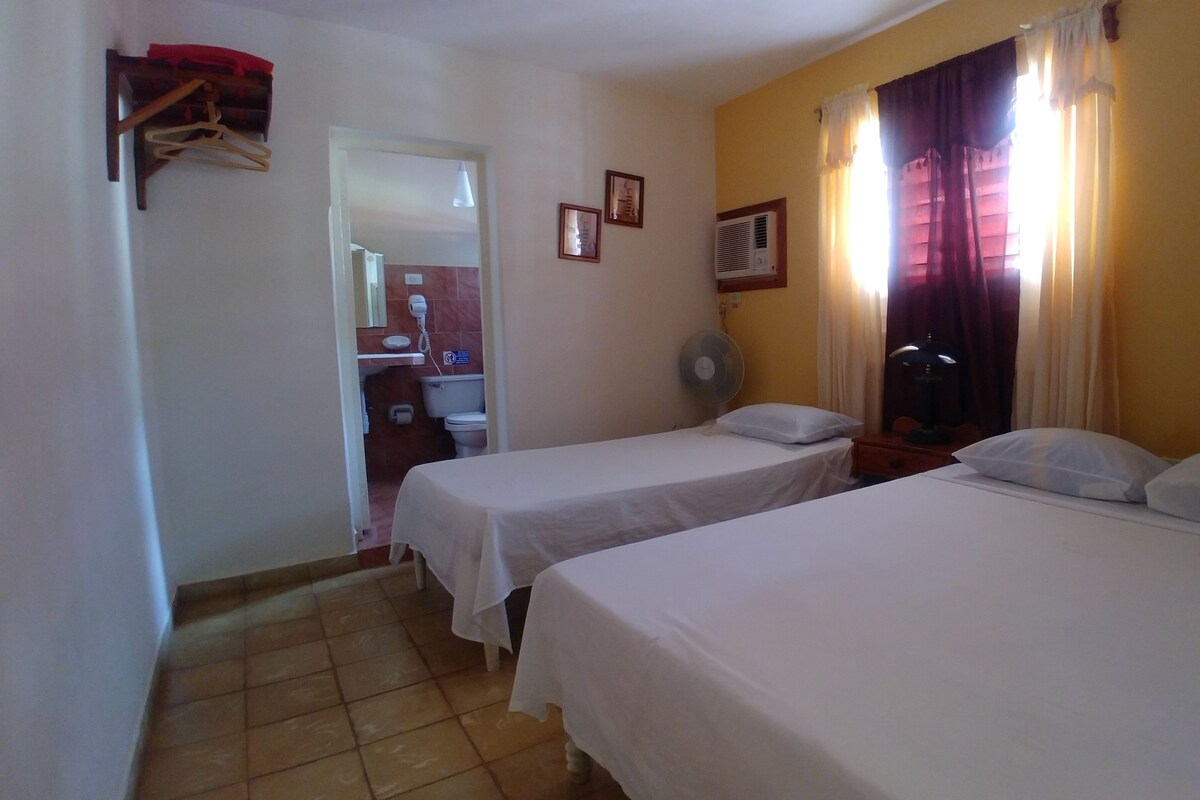 Une chambre d’hôtel avec deux lits, un ventilateur, une table de chevet et une salle de bain visible à travers une porte ouverte.
