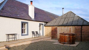 A building with a white exterior, a wooden door, and a chimney. There are two chairs outside and a wooden barrel.
