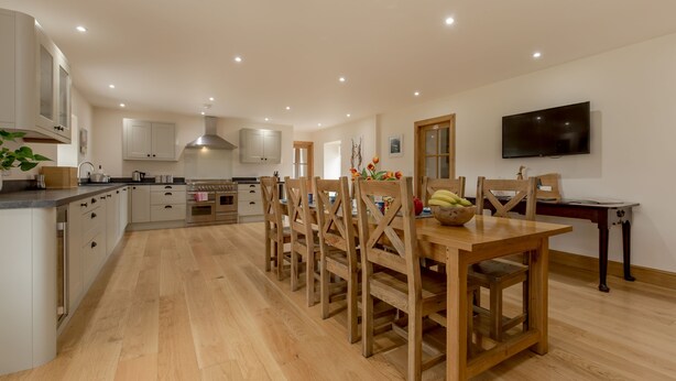 A spacious kitchen with wooden cabinets, a dining table with chairs, and a TV mounted on the wall.
