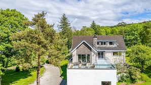 Een modern huis met een groot glazen balkon, omringd door weelderige begroeiing en bomen.
