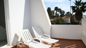 A balcony with two white lounge chairs, a tiled floor, and a view of palm trees and buildings in the background. - Oliva: House / Villa - Oliva (Oliva)
