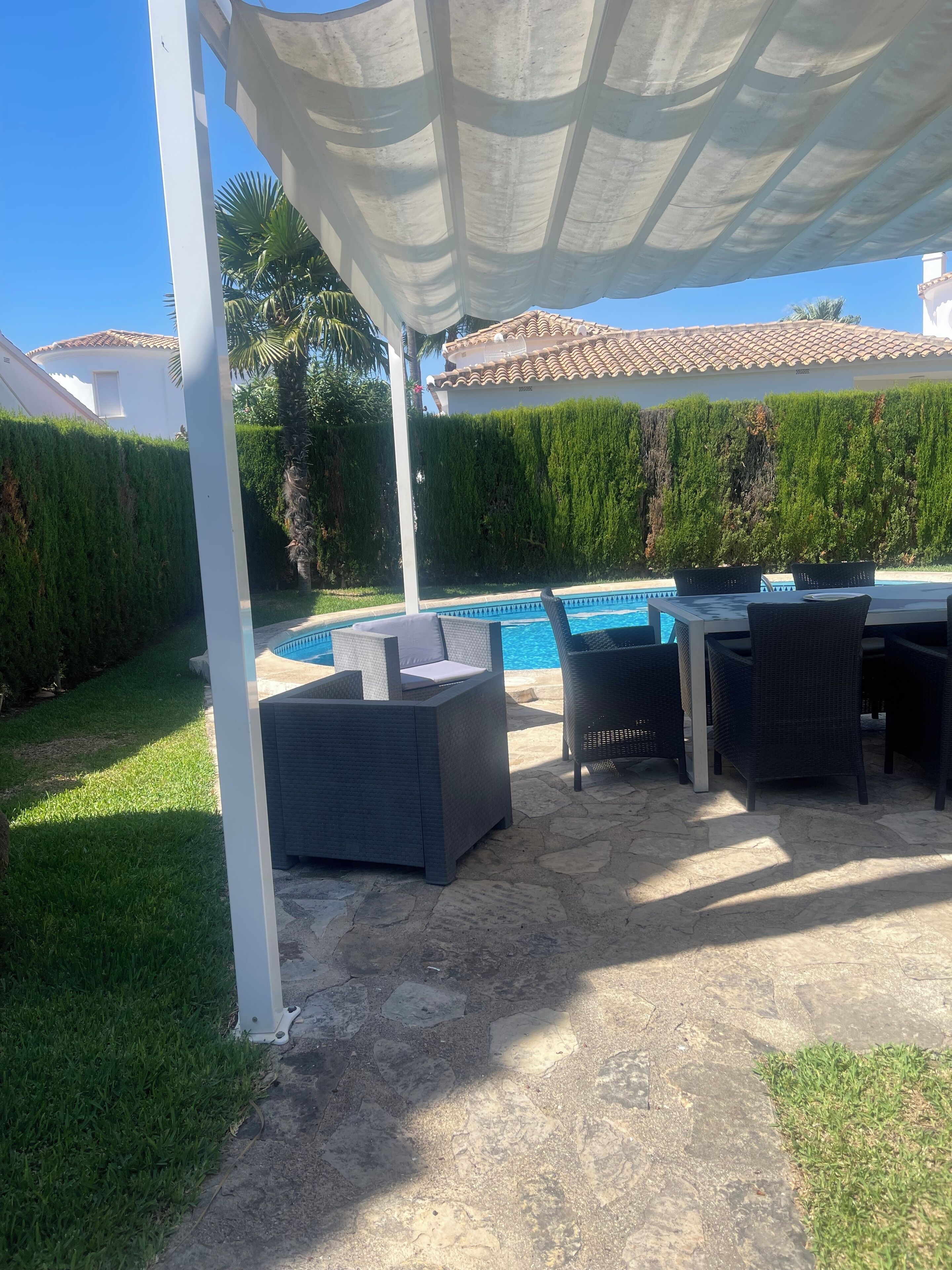 Un patio exterior con piscina, mobiliario de mimbre y una pérgola blanca que proporciona sombra.