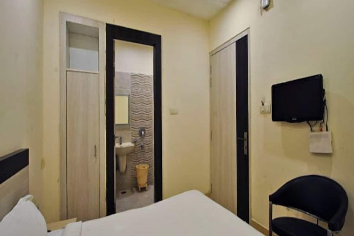 Un dormitorio con cama, una silla, televisor y baño visible a través de una puerta abierta.