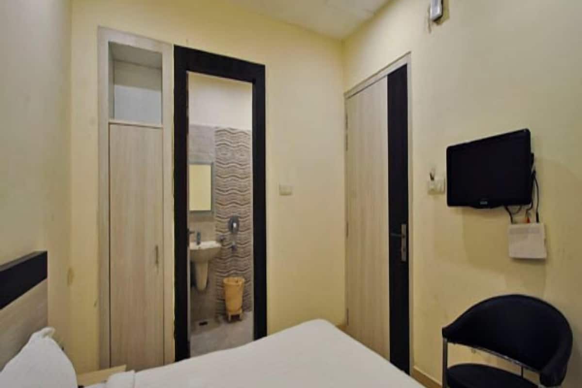 Un dormitorio con cama, una silla, televisión y un baño visible a través de una puerta abierta.