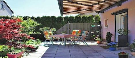 An outdoor patio with a table and chairs, surrounded by potted plants and a wooden pergola.