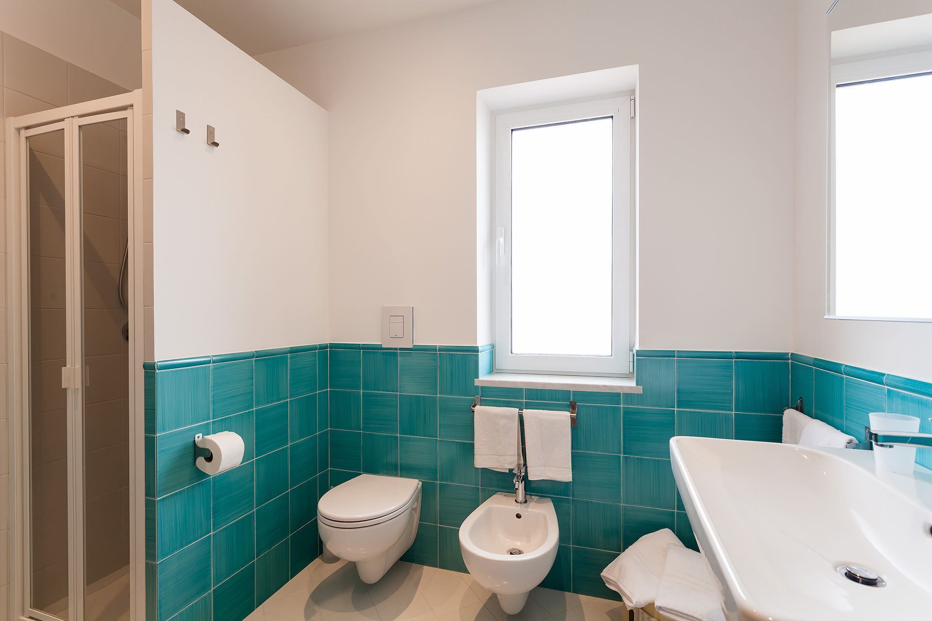 Un baño con inodoro, bidé y bañera, con azulejos verde azulado y blancos y un ventanal.