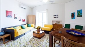 Een woonkamer met een bank, een gele fauteuil, een houten eettafel en een plafondventilator.