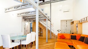 Um espaço de estar moderno com escada de madeira, área de jantar com mesa de vidro e sofá laranja.