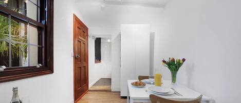 Ein schmaler Flur führt zu einem Essbereich mit einem Tisch für zwei Personen, einer Holztür und einem Fenster mit Jalousien.