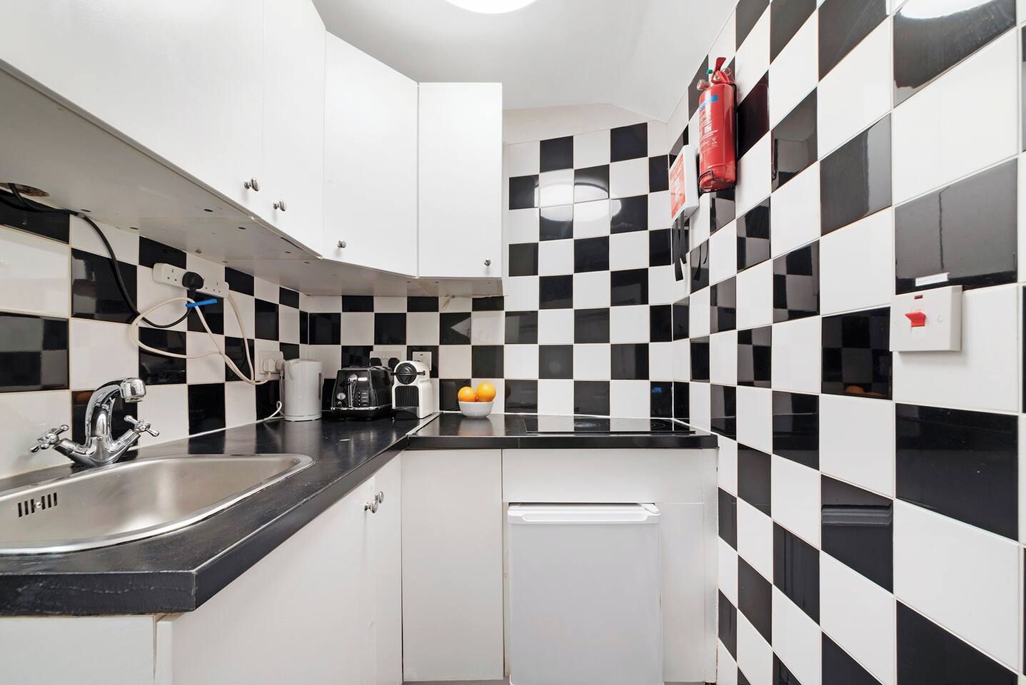 Una cocina con salpicadero blanco y negro a cuadros, electrodomésticos de acero inoxidable y un extintor de incendios rojo montado en la pared.