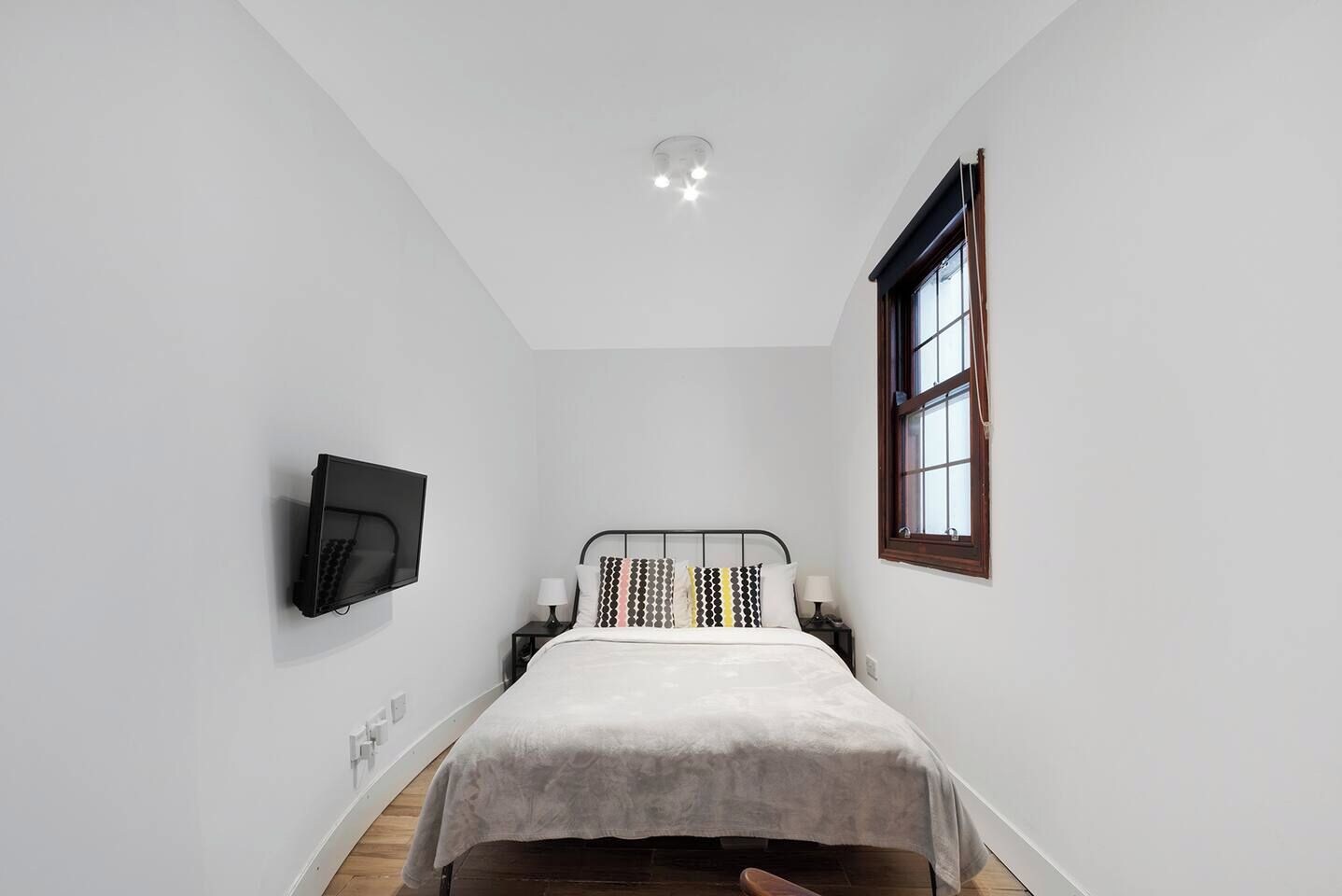 A bedroom with a bed, a television mounted on the wall, a window, and a ceiling light.
