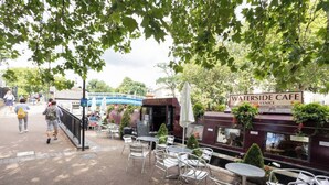 Een terras met tafels en stoelen, een bordje met 'Waterside Cafe' en een pad met mensen.