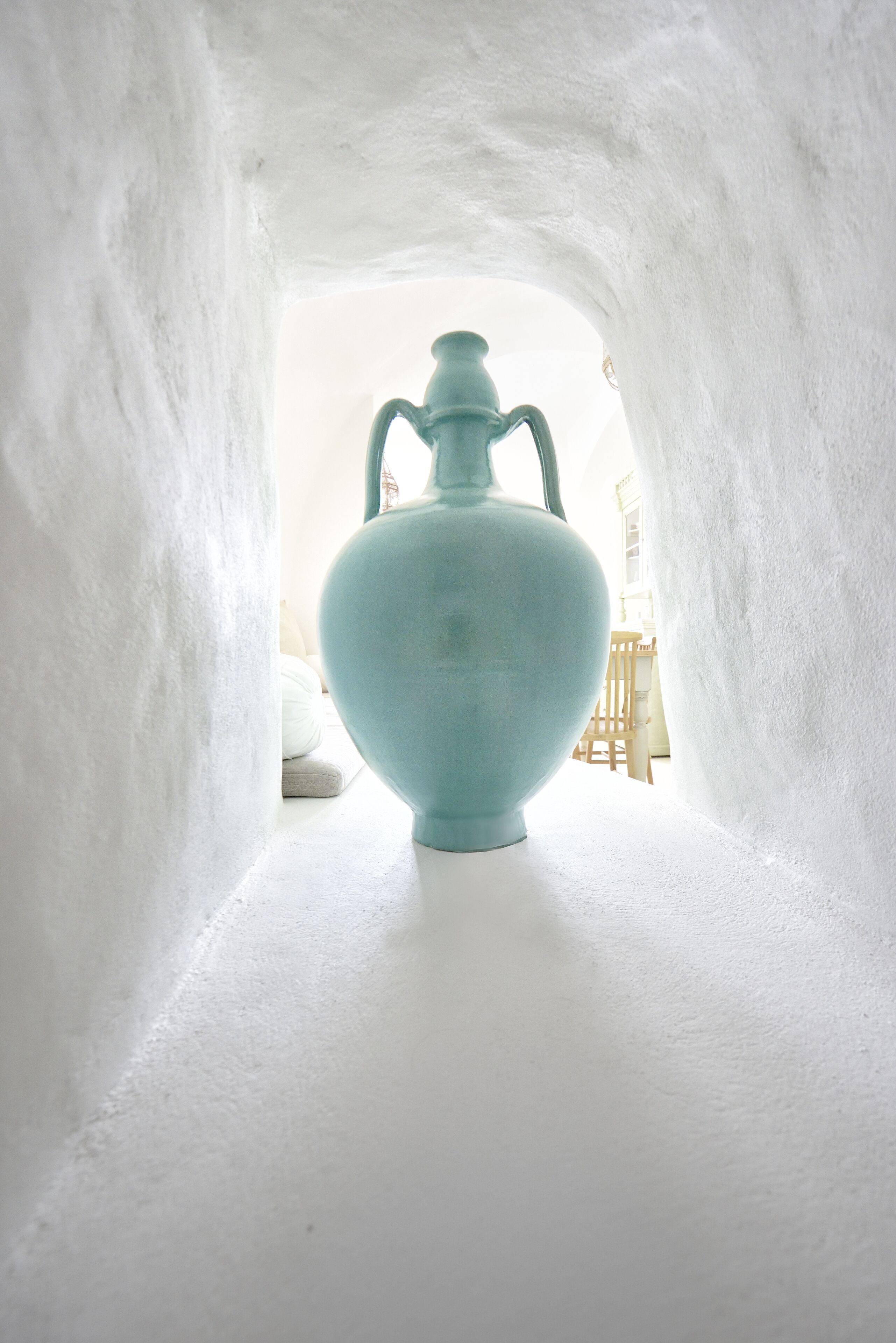 Eine hellblaue Keramikvase steht in einer weißen, gewölbten Nische. Im Hintergrund sind ein Holzstuhl und ein weißer Tisch zu sehen.