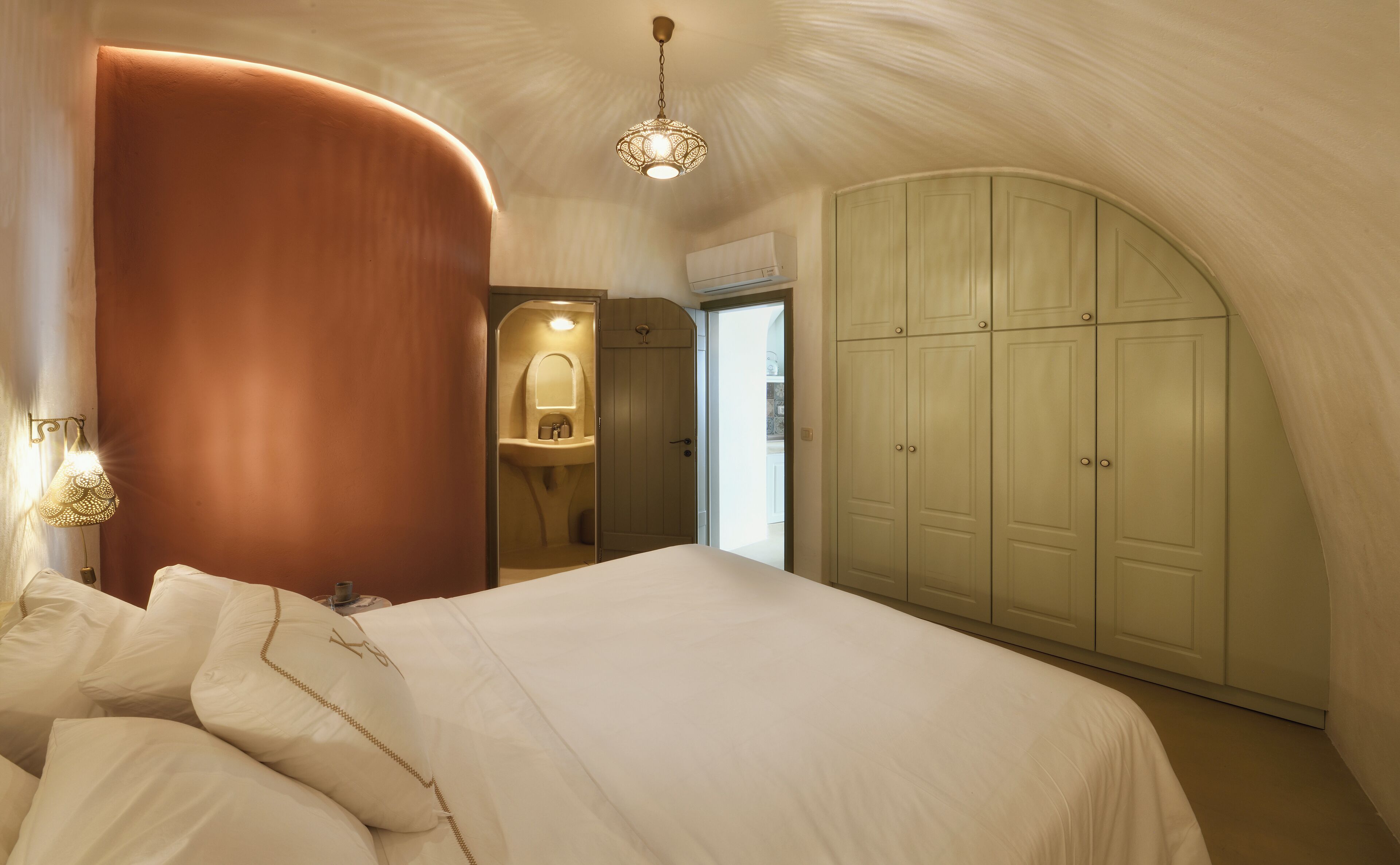 A bedroom with a curved wall, a bed with white bedding, a hanging light fixture, and a wardrobe.