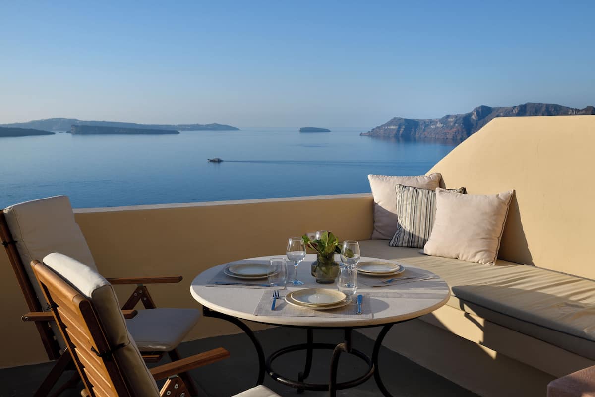 A round table set with plates, glasses, and cutlery on a balcony overlooking a calm sea with distant islands.