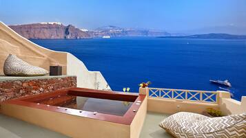 A sunlit terrace with a hot tub, stone wall, and a view of the sea and cliffs.