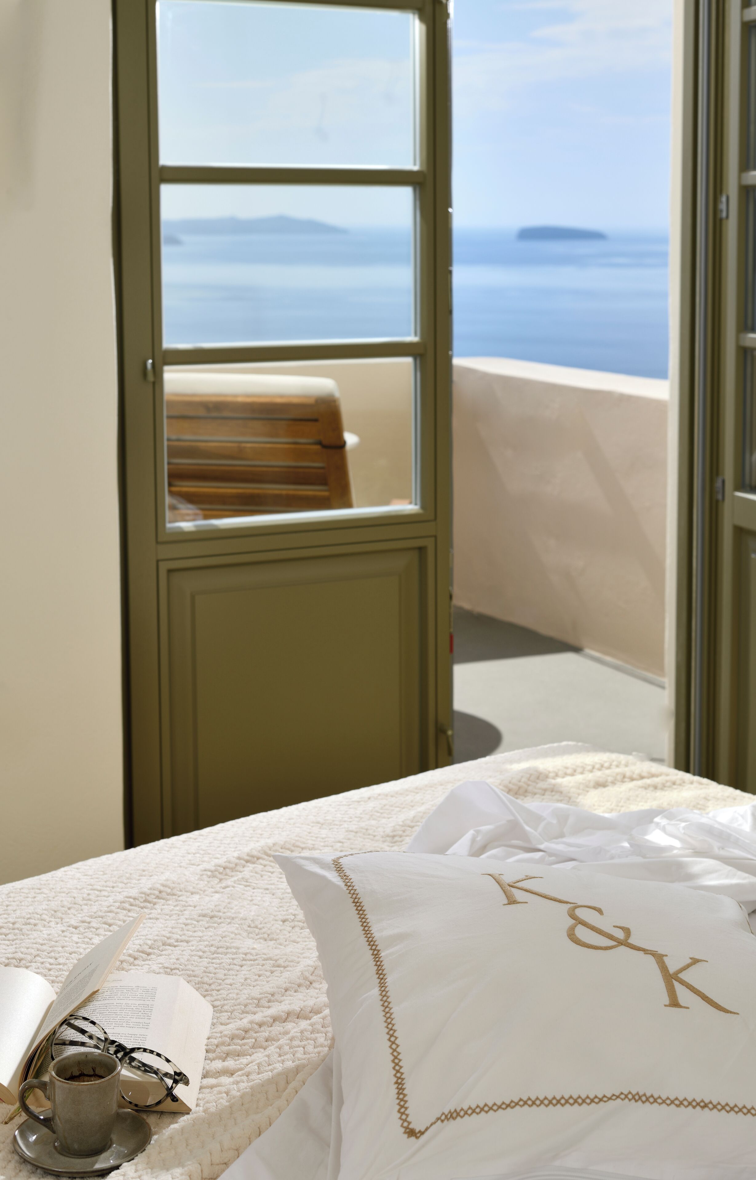 Habitación de hotel con balcón, cama con edredón blanco, almohada con iniciales, taza y libro abierto.
