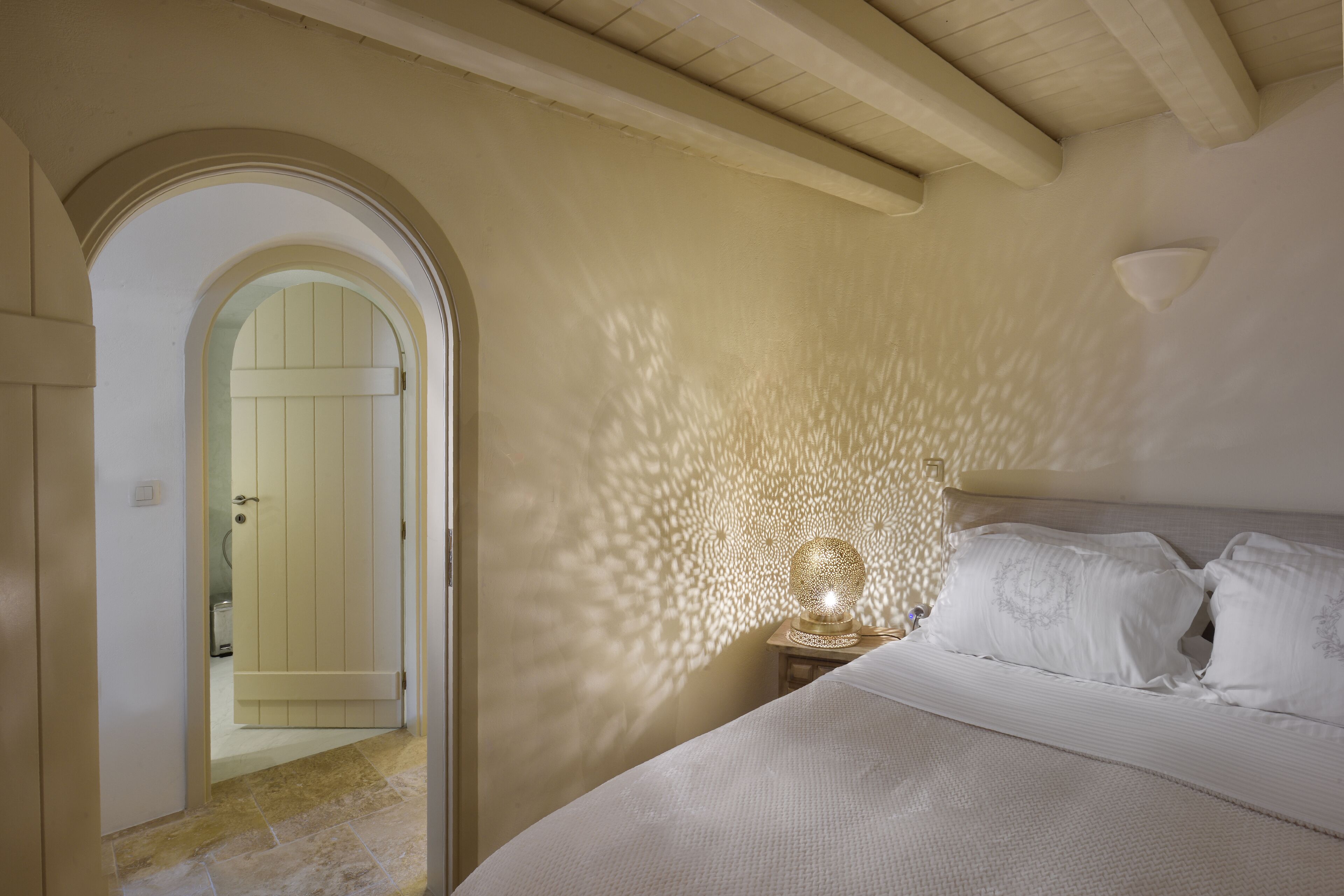 Un dormitorio con una cama, una mesita de noche, una lámpara de pared y un arco que da paso a otra habitación.