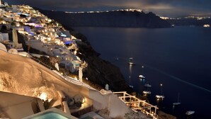 A rooftop terrace with a hot tub, seating area, and a view of a coastal town at night. - The A Collection, Oia by K&K (Santorini)