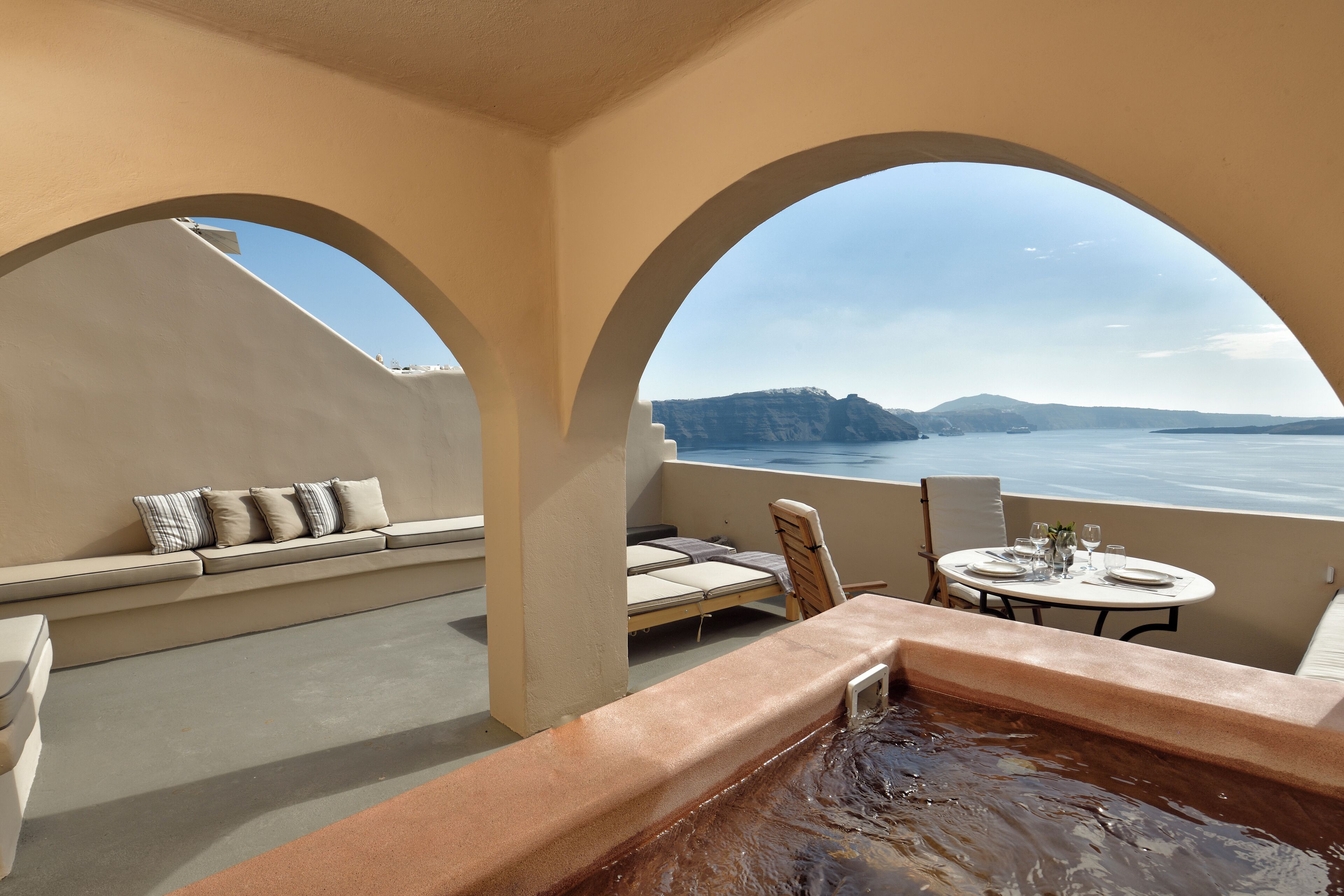 A rooftop terrace with a hot tub, seating area, and a table set for dining, overlooking a body of water and distant land.