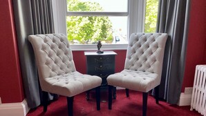 A room with two tufted armchairs, a small table, and a view of a street through a window with curtains. - Blenheim Guest House York (York)