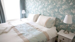 A neatly made bed with a light-colored bedspread and pillows, a bedside table with a lamp, and a floral-patterned wall. - Smithfield guesthouse (Dounby)