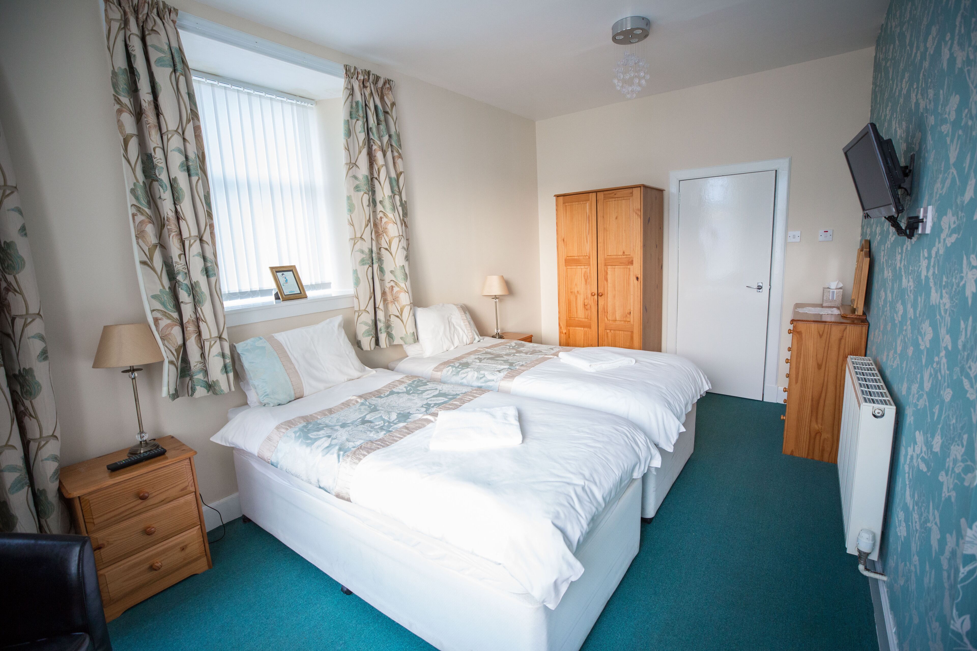 A bedroom with two beds, a wooden wardrobe, a radiator, a TV, and a window with curtains.