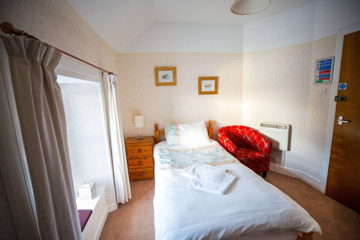 A hotel room with a bed, a bedside table, a chair, a window with curtains, and a door.