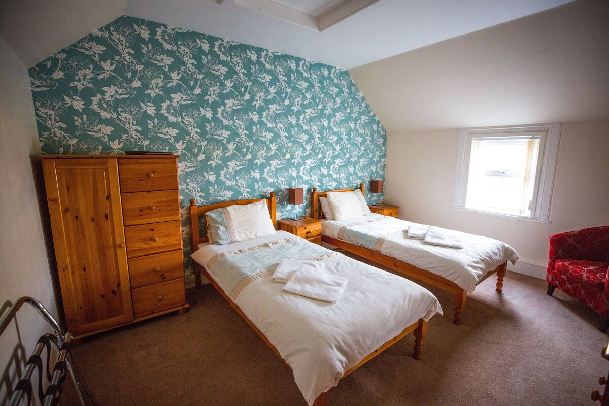 A bedroom with two single beds, a wooden wardrobe, a red armchair, and a window with curtains.