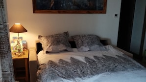 A bedroom with a large bed, a nightstand with a lamp, a painting on the wall, and a door with a heart-shaped decoration.
