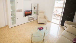 Una sala de estar moderna con una mesa de centro de vidrio, un sillón color beige y un televisor de pantalla plana sobre un mueble blanco.