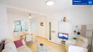 Una sala moderna con un sofá, sillones, una mesa de centro de vidrio, un televisor y una zona de comedor con una mesa y sillas.