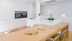Cocina moderna con armarios blancos, una isla central y un comedor con mesa de madera y sillas doradas.