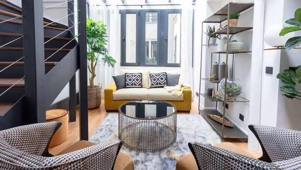Una sala de estar moderna con una escalera, un sofá amarillo, una mesa de centro y una estantería con plantas.