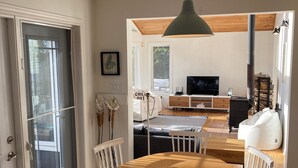 Ein Essbereich mit Holztisch und weißen Stühlen, einer hängenden Pendelleuchte und Blick in den Küchenbereich mit Fernseher und Kaminofen.