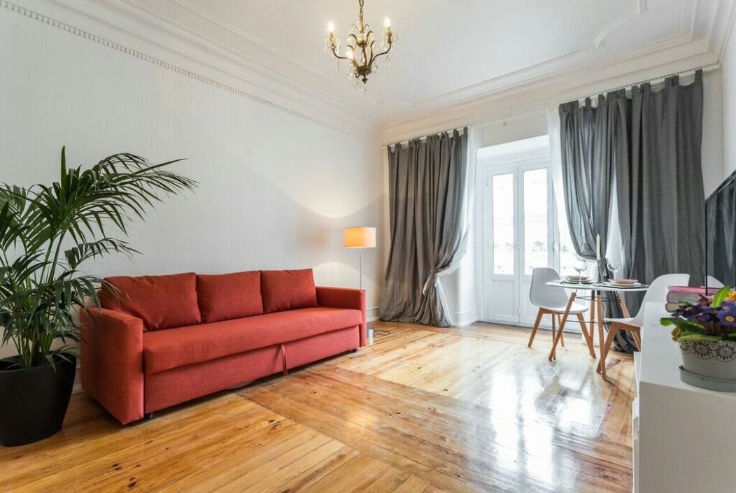 A room with a red sofa, wooden flooring, a potted plant, a table with chairs, and a window with curtains.