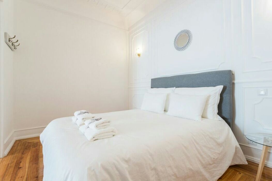 A neatly made bed with white linens and towels, a round mirror on the wall, and a wooden floor.