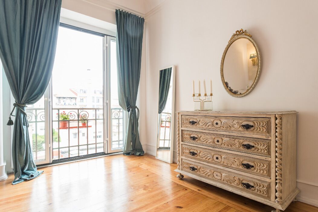 A room with a wooden floor, a white dresser with ornate handles, a large mirror, and blue curtains.