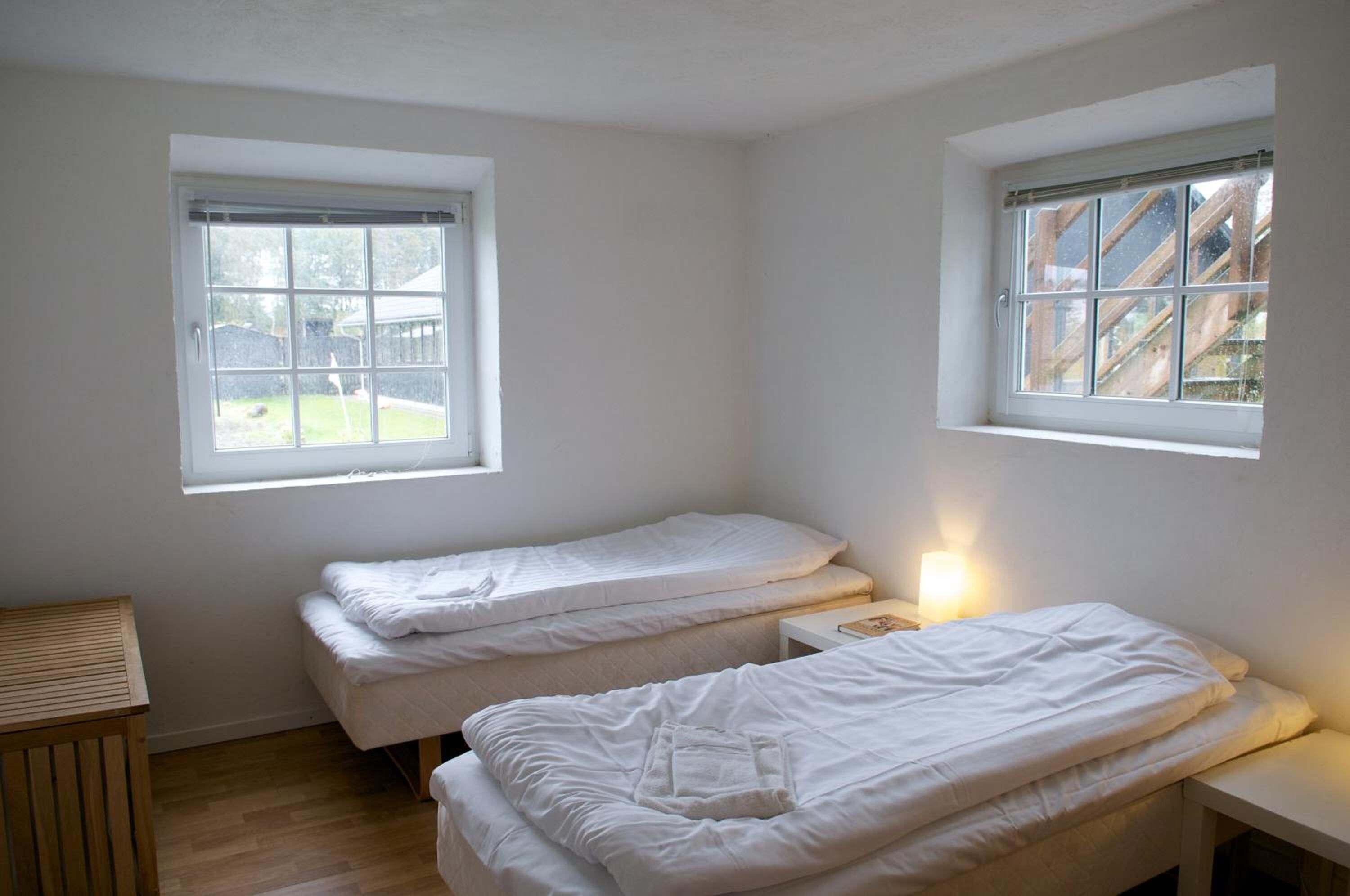 A small, clean room with two single beds, a small table, and two windows providing natural light.