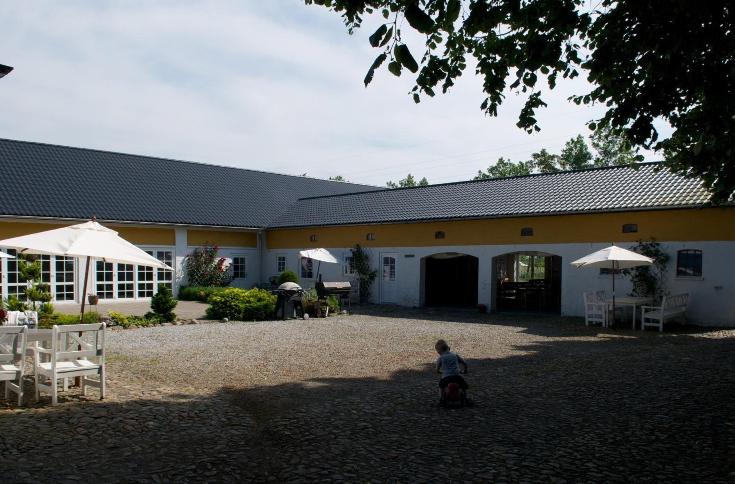 Een erf met een geplaveide vloer, een geel gebouw met meerdere ramen, en buiten zitplaatsen met parasols.