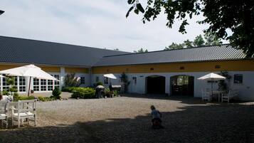 Een erf met een geplaveide vloer, een geel gebouw met meerdere ramen, en buiten zitplaatsen met parasols.